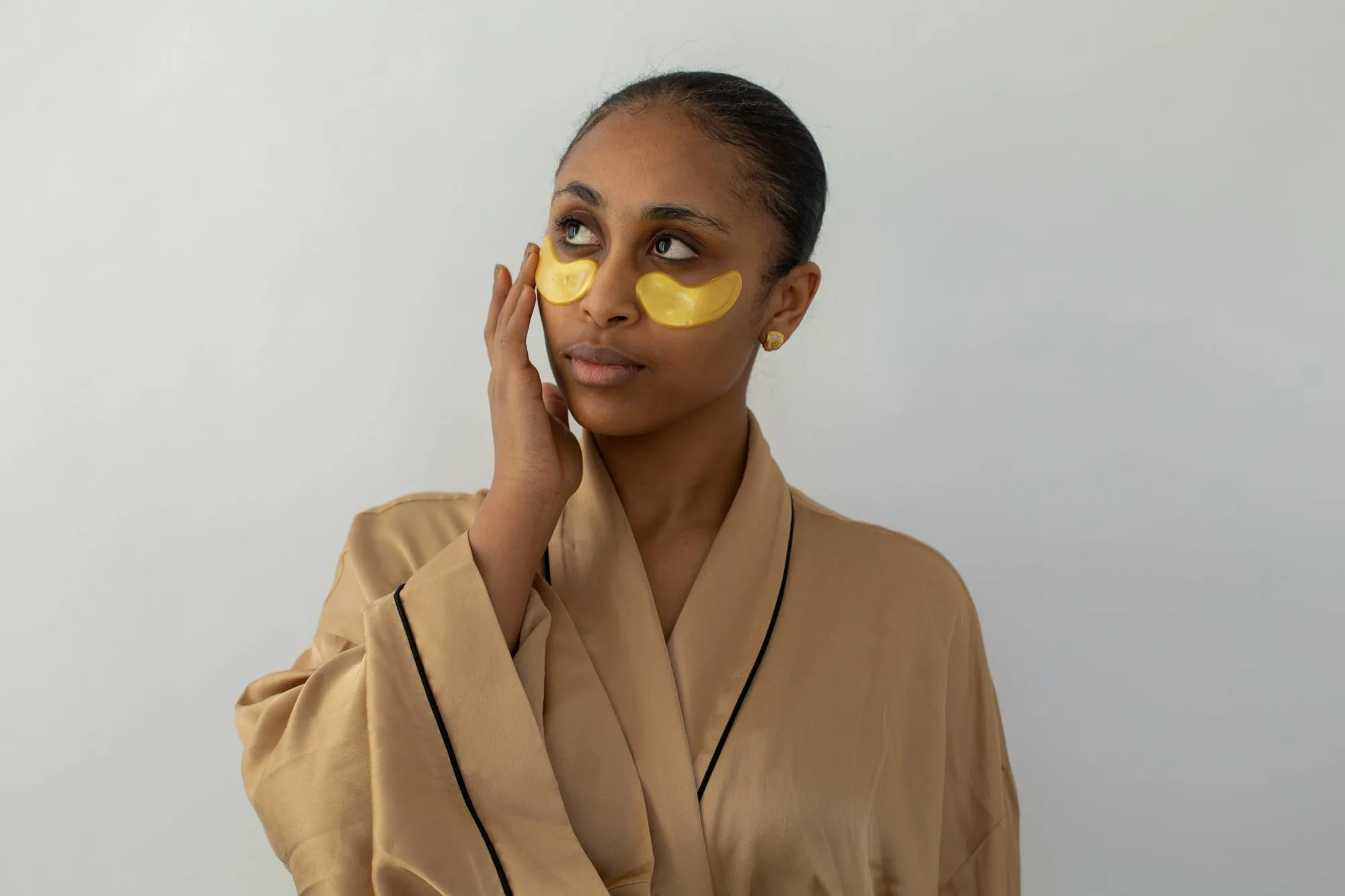 Woman wearing gold under-eye patches in a robe, promoting skincare and relaxation. Editorial photograph for our Beauty Eyes guide for Dubai shoppers.